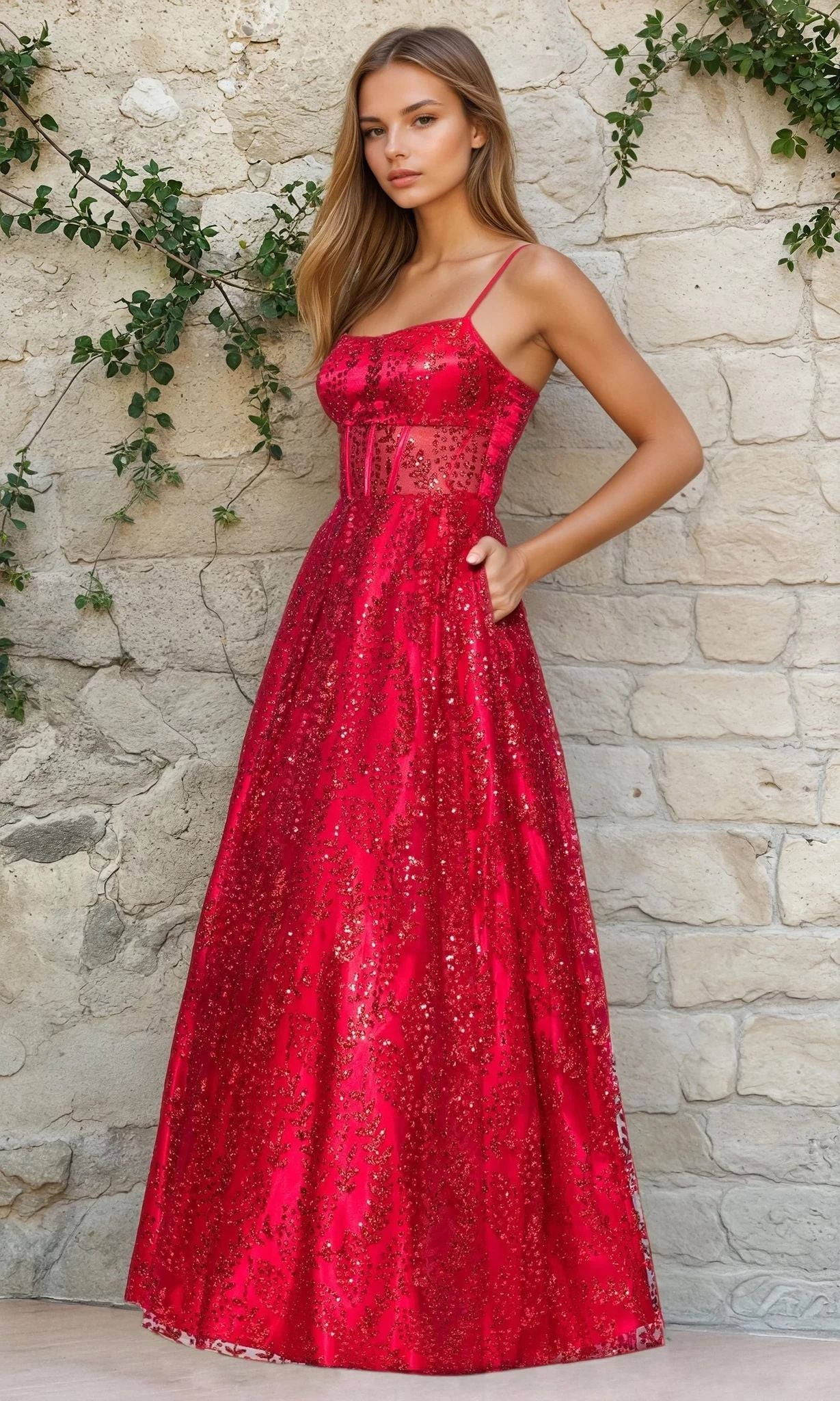 A woman in a red Glitter-Print Prom Ball Gown (Blondie Nites 5539BN) with thin straps and a subtle corset bodice stands against a light stone wall decorated with green vines.